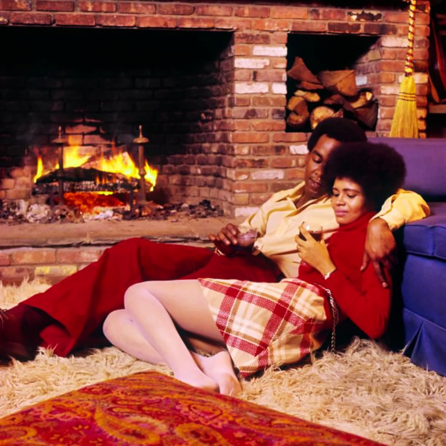 A couple relaxes on a shaggy carpet in front of a brick fireplace, holding drinks. A fire burns behind them, and stacked firewood is visible. The atmosphere is warm and cozy.