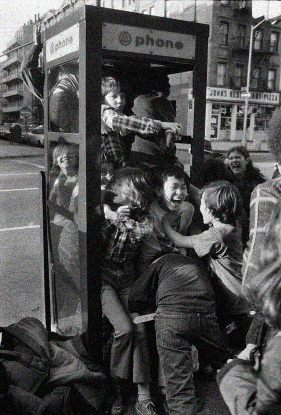 A group of laughing children squeeze into and around a phone booth on a city street, with some climbing inside and others pushing to fit in, creating a lively and joyful scene.