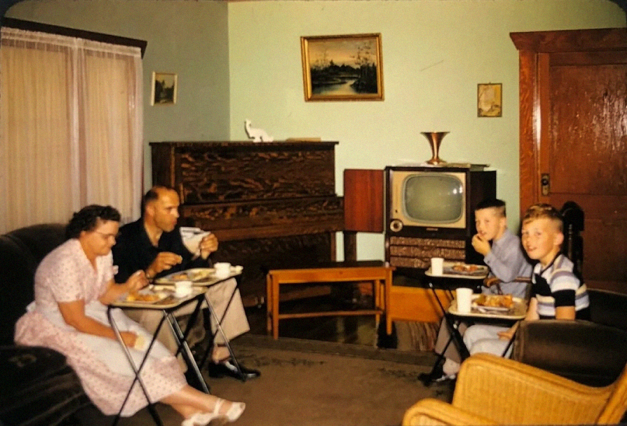 A family of four eats dinner on TV trays in a vintage living room, with an old-fashioned television, upright piano, and paintings on the walls. The parents sit on a sofa, while two boys sit on chairs, smiling.