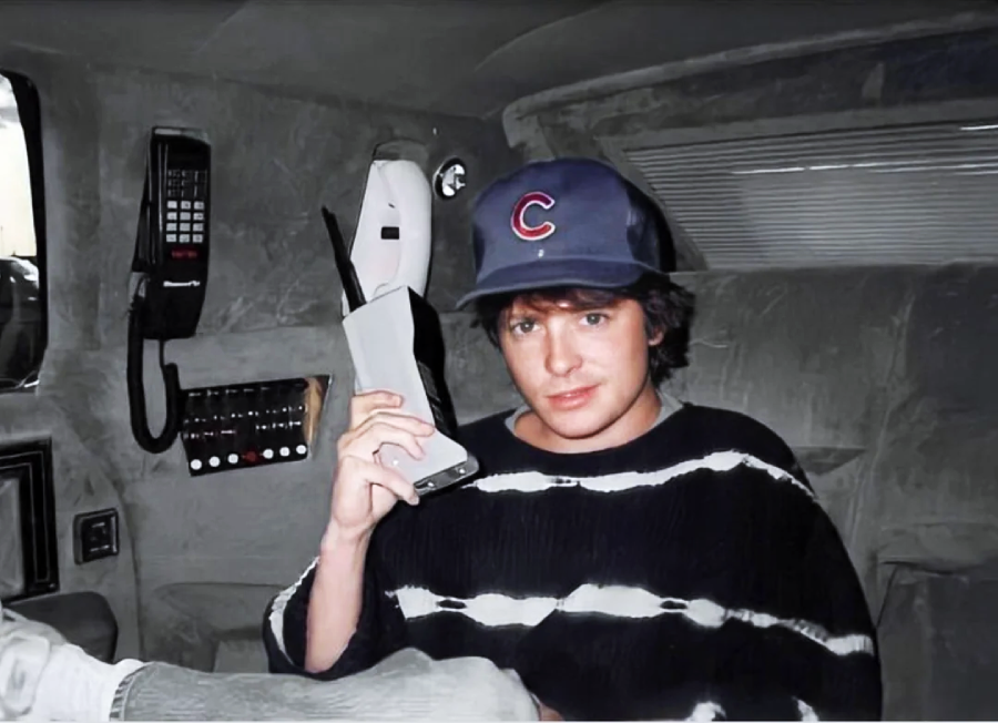 A young person in a striped shirt and Chicago Cubs baseball cap sits in a plush car interior, holding a large vintage mobile phone to their ear, with another phone and control panel visible beside them.