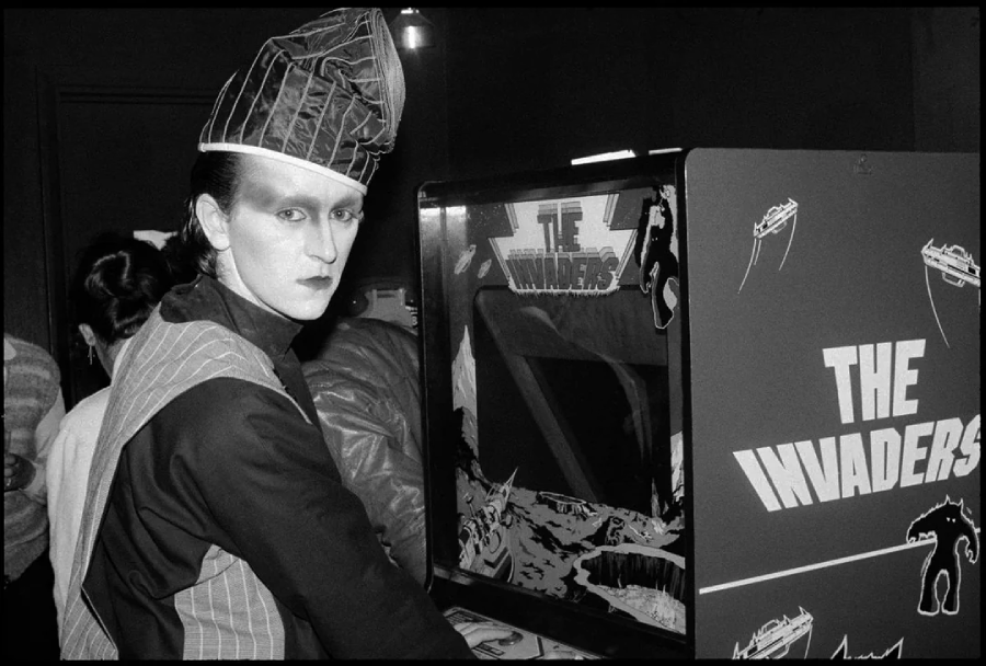 A person in dramatic makeup and a tall, angular hat stands next to a vintage arcade machine labeled "THE INVADERS," turning toward the camera with a serious expression in a dimly lit room.