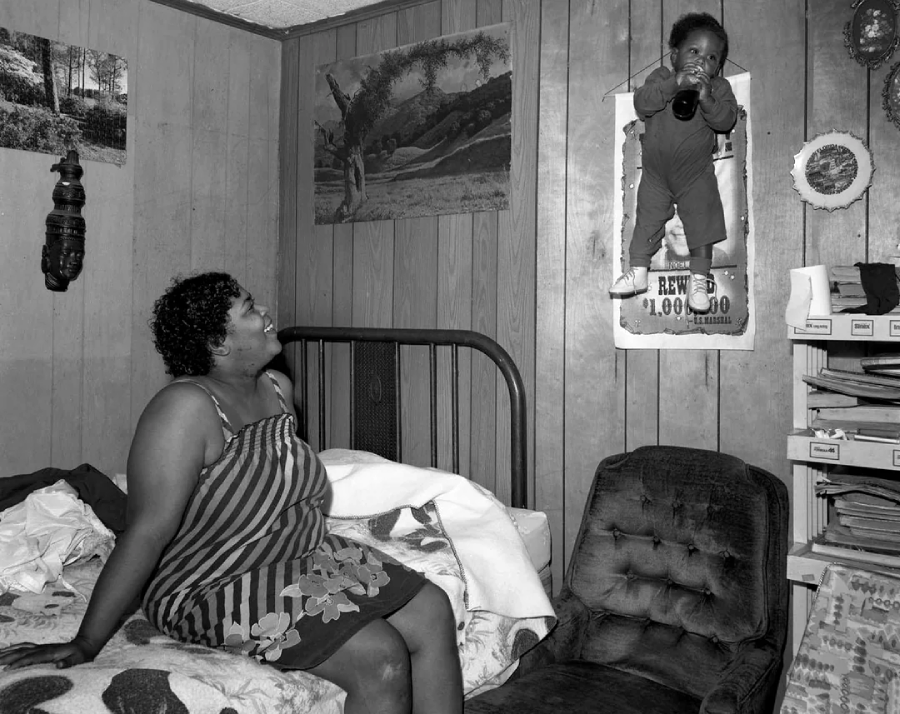 A woman sits on a bed in a wood-paneled room, smiling up at a baby who is standing on a wall-mounted calendar, holding onto a bottle. The room has various wall decorations, a chair, and stacks of papers.