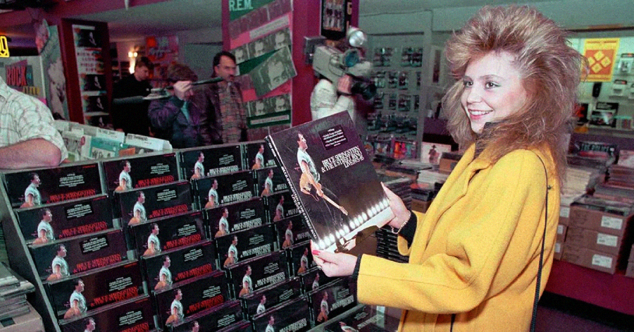 A woman in a yellow coat smiles while holding a Bruce Springsteen record in a busy record store, surrounded by stacks of the same album and people browsing in the background.