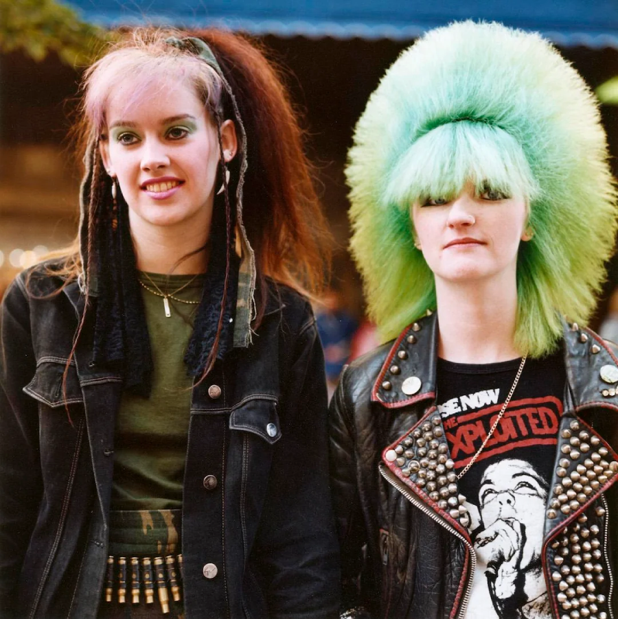 Two young women with punk hairstyles and fashion stand together. One has long dark hair with colored streaks; the other has a large, bright green mohawk. Both wear dark jackets, one with metal studs and band patches.