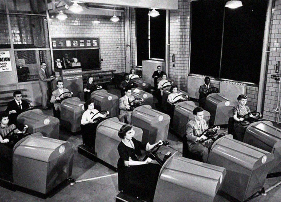 A black-and-white photo shows men and women sitting in rows of vintage driving simulators in a classroom, each holding a steering wheel, while an instructor observes from the front.