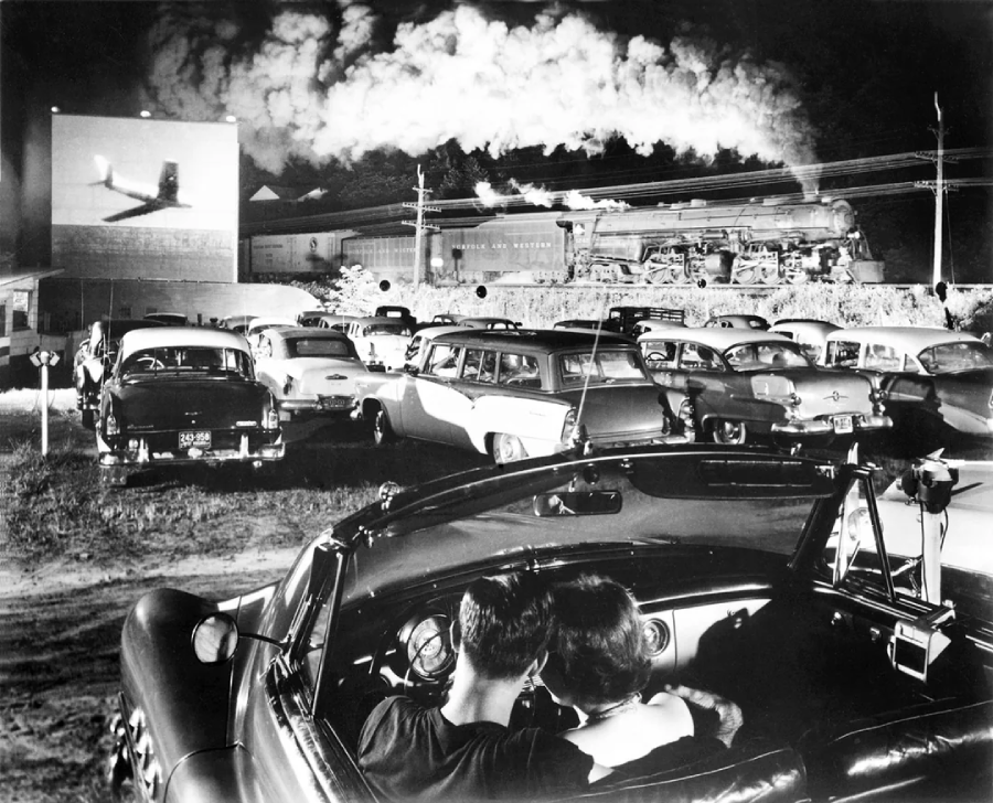 A black-and-white photo of a crowded drive-in theater, showing couples in classic cars watching a film as a steam train passes by in the background, with smoke filling the night sky.