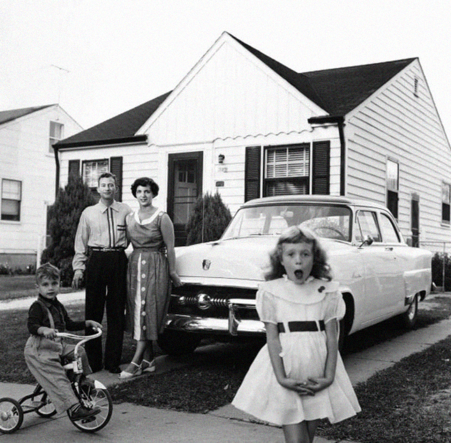 A vintage black-and-white photo of a family in front of a classic car and a house. The parents pose together, a boy sits on a tricycle, and a surprised girl stands in front, all dressed in 1950s-style clothing.