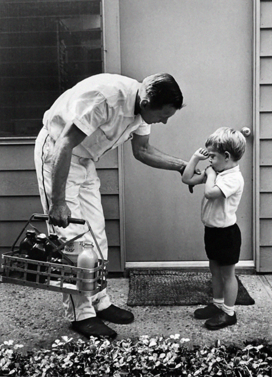 A milkman in a white uniform gently shows a young boy how to flex his arm. The boy stands shyly with hands near his mouth as the milkman holds a crate of bottles outside a house.