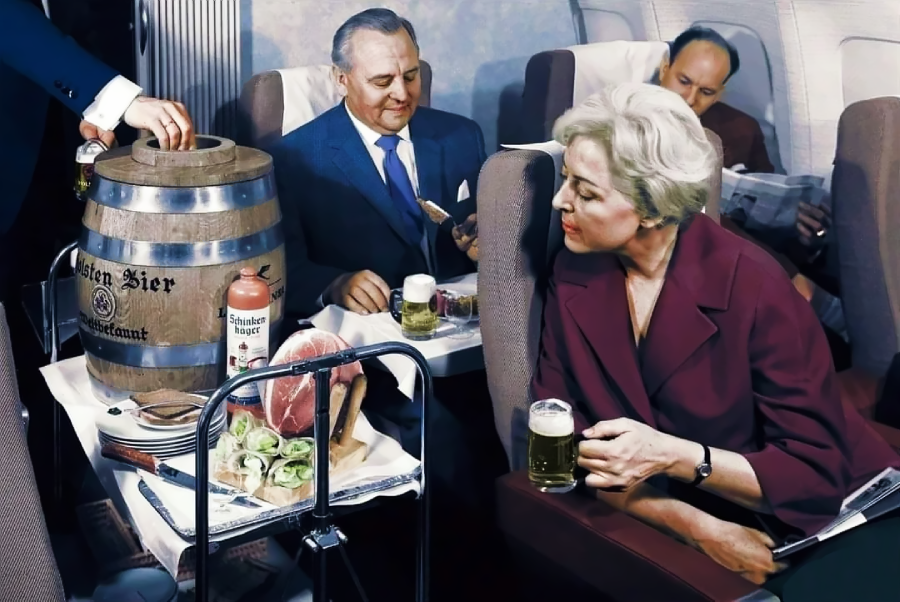Passengers on an airplane are served beer from a wooden keg along with food. A woman in front holds a glass of beer, while two men behind her enjoy drinks and snacks in a vintage, luxurious airline cabin setting.