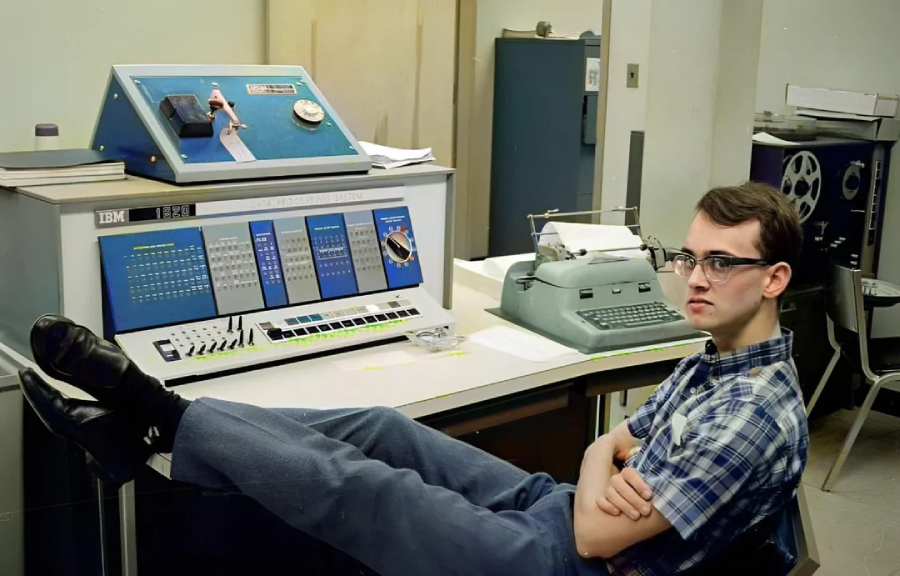 A young man in glasses sits with his feet up on a desk in front of a vintage IBM computer, looking at the camera. Nearby are a typewriter and various office equipment in a retro-style room.