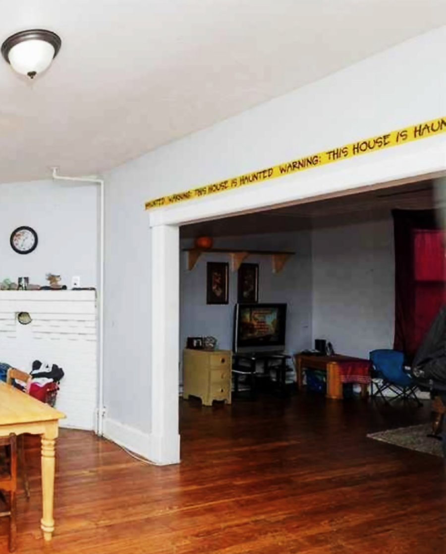 A living room with hardwood floors, a dining table on the left, a white brick fireplace, and a TV. A yellow banner reading "WARNING: THIS HOUSE IS HAUNTED" hangs above the doorway to a darker adjacent room.