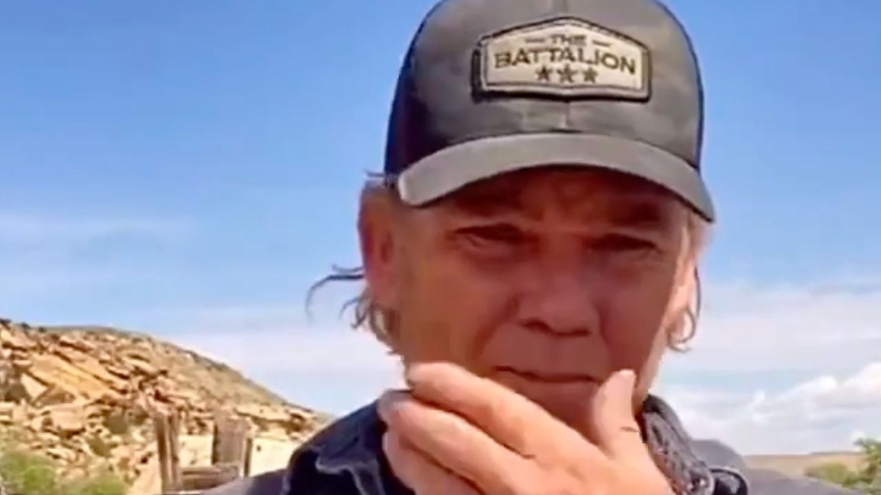 A person wearing a black cap with “THE BATTALION” and three stars on it stands outdoors, touching their chin, with a blue sky and rocky landscape in the background.