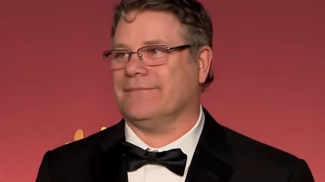A man in glasses and a black tuxedo with a bow tie stands in front of a red backdrop, smiling slightly.