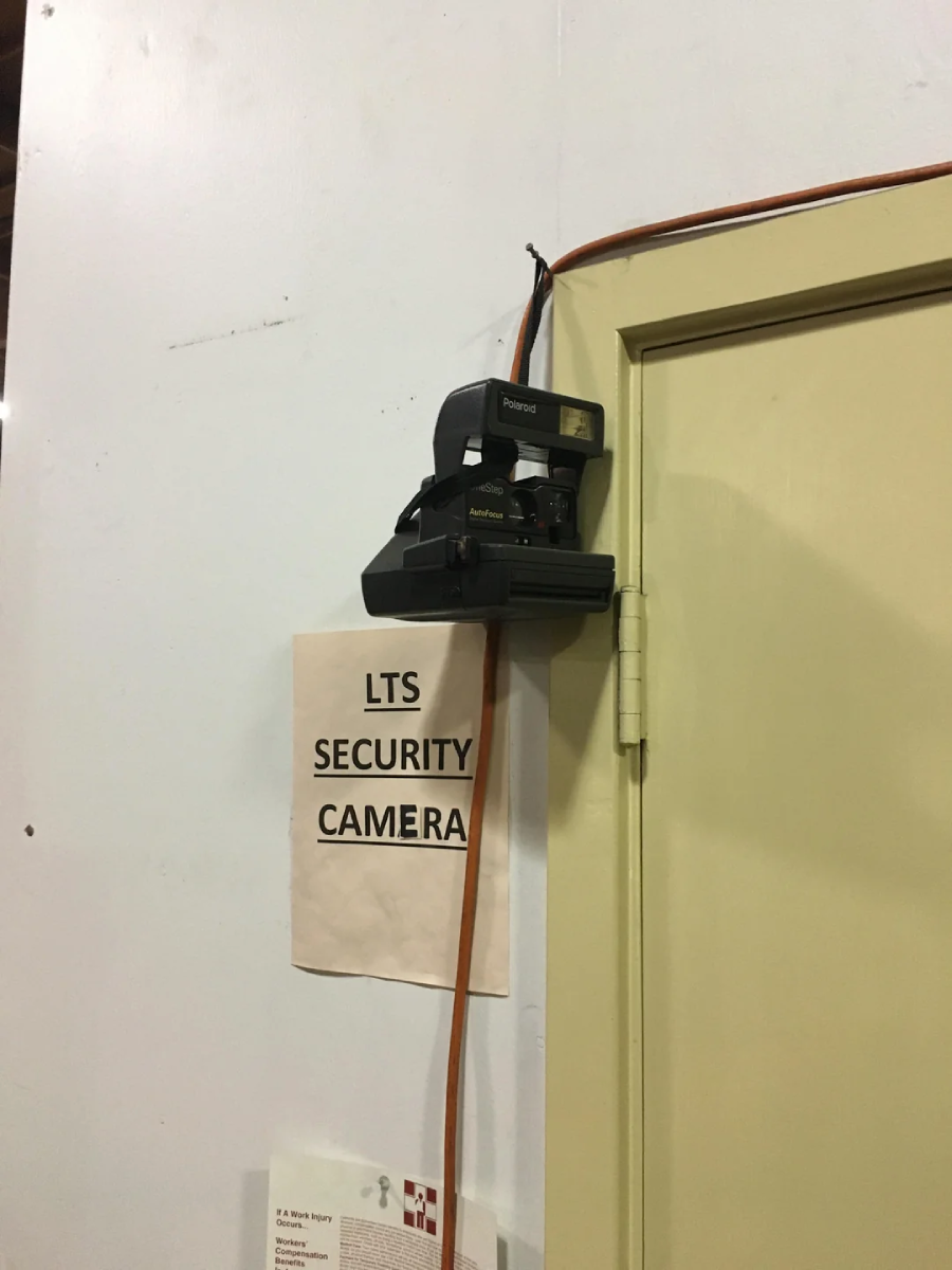 A Polaroid instant camera is mounted above a door with an orange cable running to it. A sign below reads "LTS SECURITY CAMERA." The setup appears humorous and makeshift.