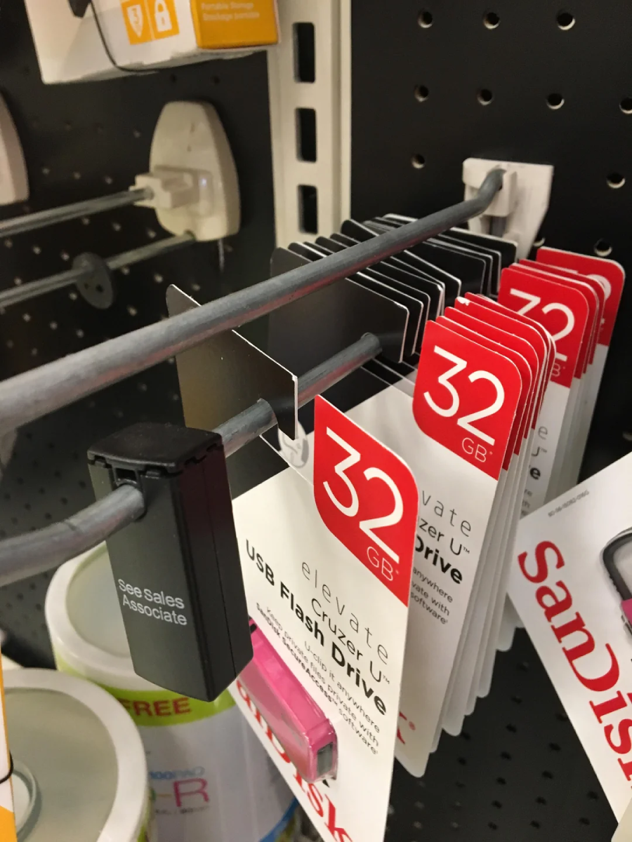 Several 32GB USB flash drives hang on display hooks in a store, each secured by a large black security tag labeled "See Sales Associate." A few other electronics are visible nearby.