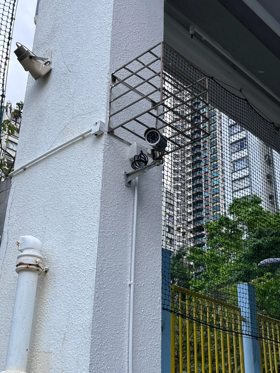 A security camera mounted on a white column with metal grating above it, next to a yellow gate and a tall building in the background. Trees and another surveillance light are also visible.