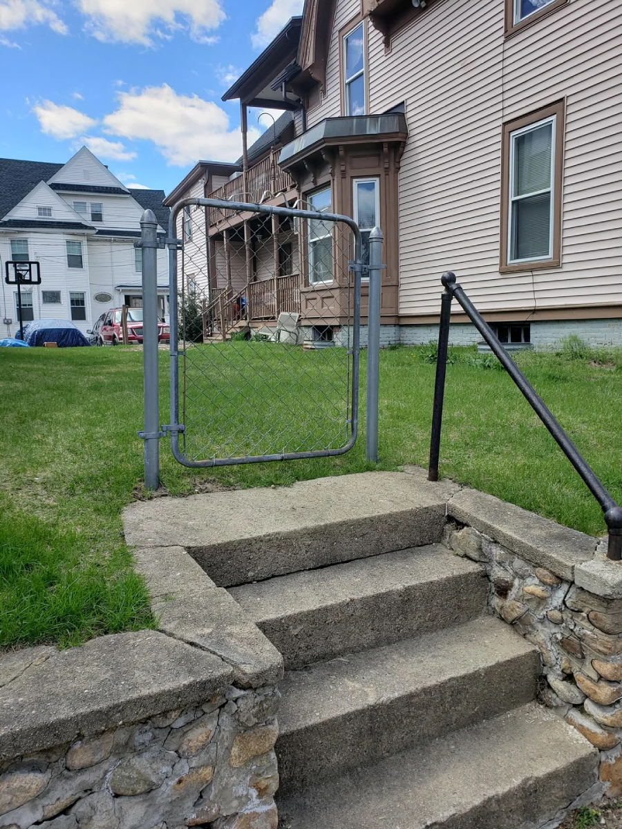 A metal gate stands at the top of a short stone and concrete stairway, but there is no fence on either side of the gate. Behind the gate is a grassy yard and a tan house with brown trim.