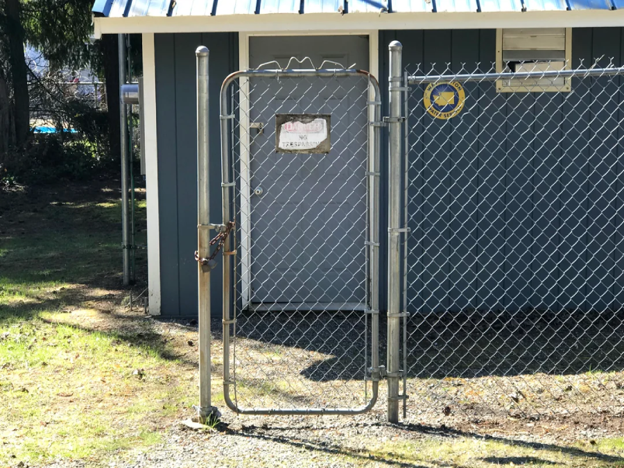A locked metal gate is installed in a chain-link fence, but the gate is not connected to the fence itself. Behind the gate is a blue building with a door and a sign. Sunlight casts shadows on the ground.