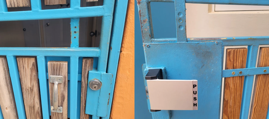 Close-up of a blue metal gate with vertical wooden panels. The gate has a handle and a lock, and a white push bar installed on it. The background shows an orange wall and part of a building.