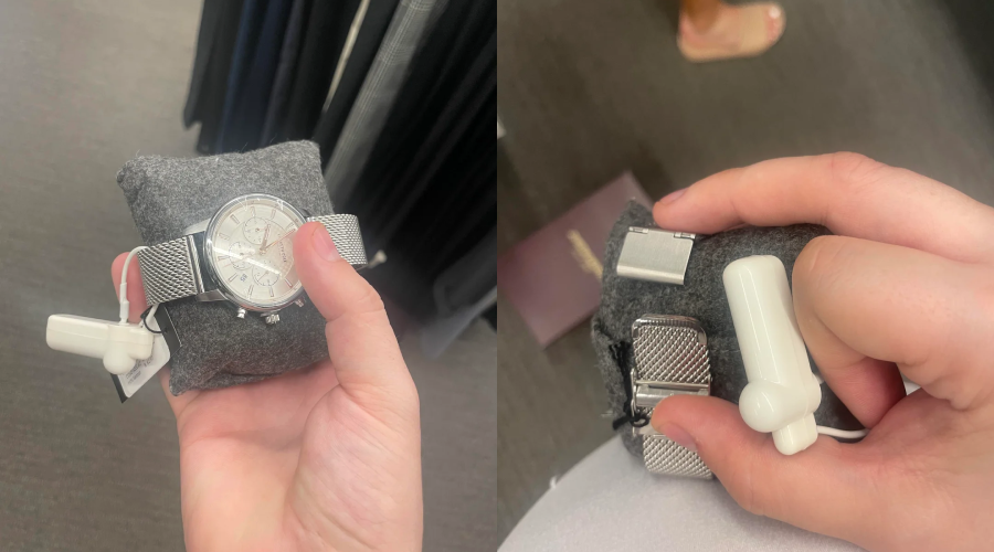 A hand holds a silver wristwatch with a mesh band and a white security tag, displayed on a gray cushion. The background is a store with gray flooring and clothing racks.