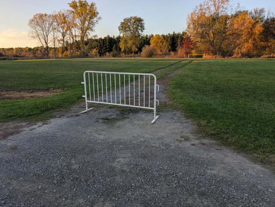 A white metal barrier blocks a gravel path leading through a grassy field with trees in the background, some showing autumn colors under a clear sky.