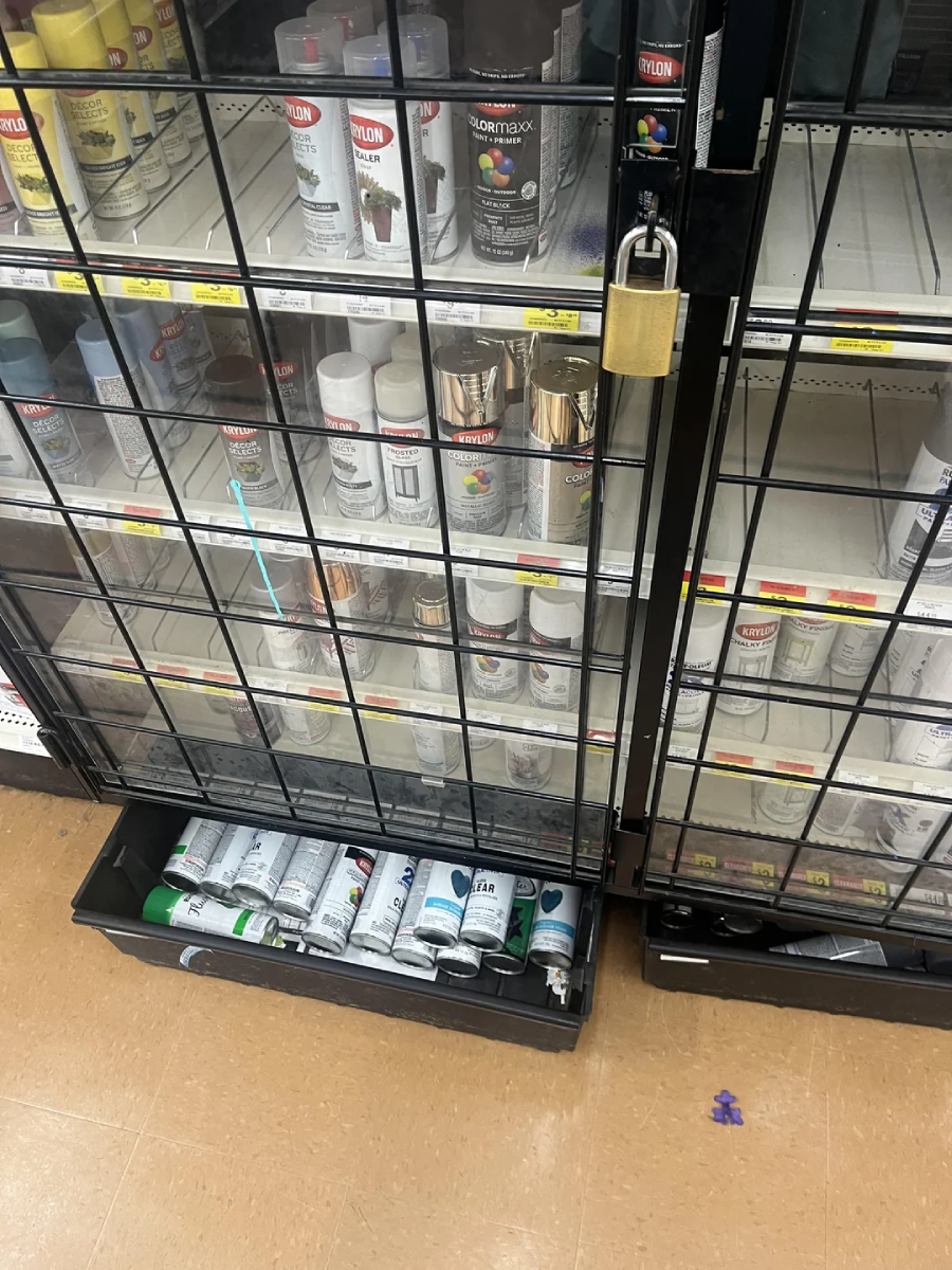 Cans of spray paint are displayed behind a locked black metal gate in a store aisle. Some cans are on the shelves, while others are in a bin at the bottom. A large gold padlock secures the gate.