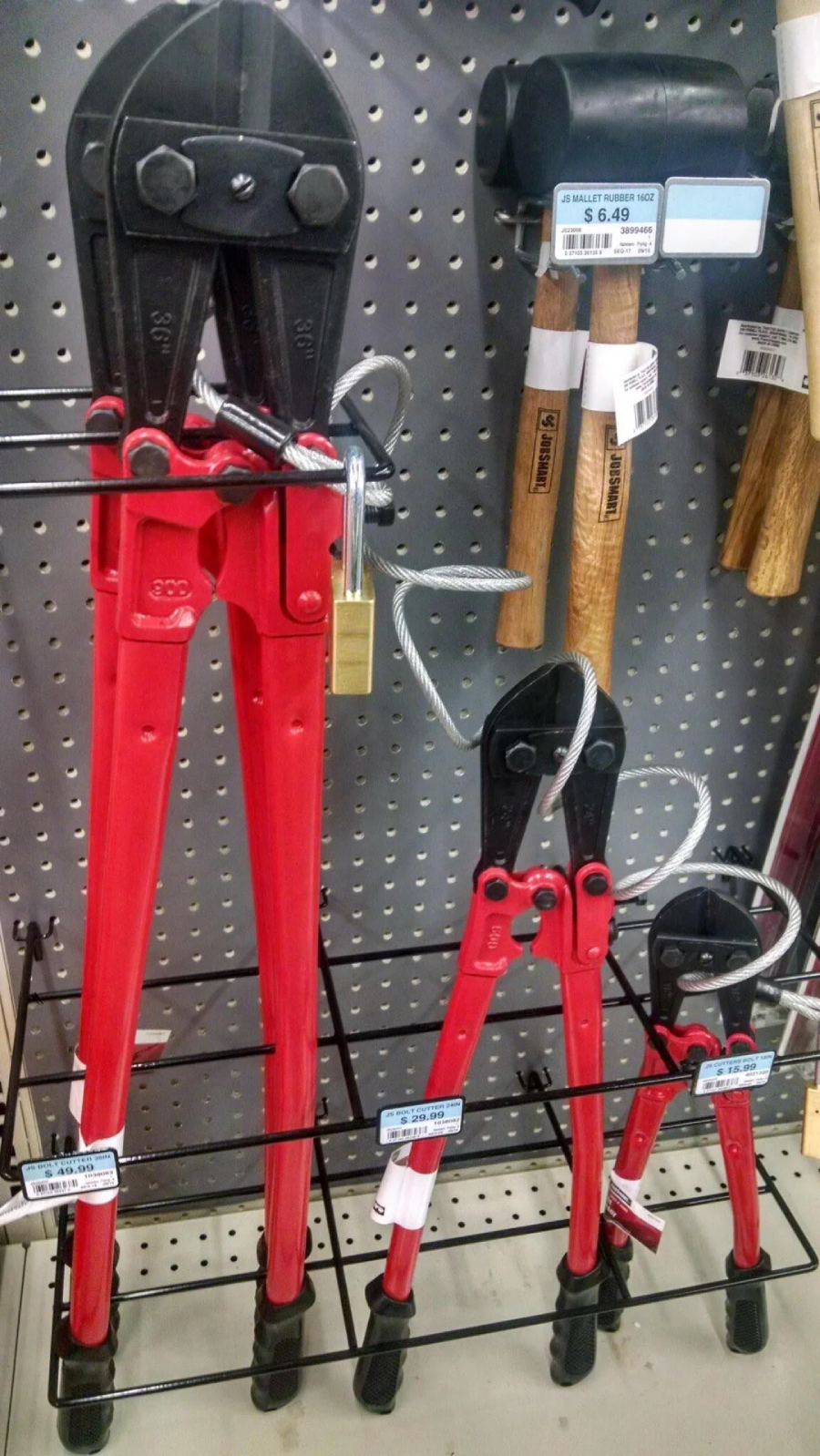 Three red-handled bolt cutters are secured by a cable and padlock on a display rack, with hammers hanging nearby on a pegboard in a hardware store.