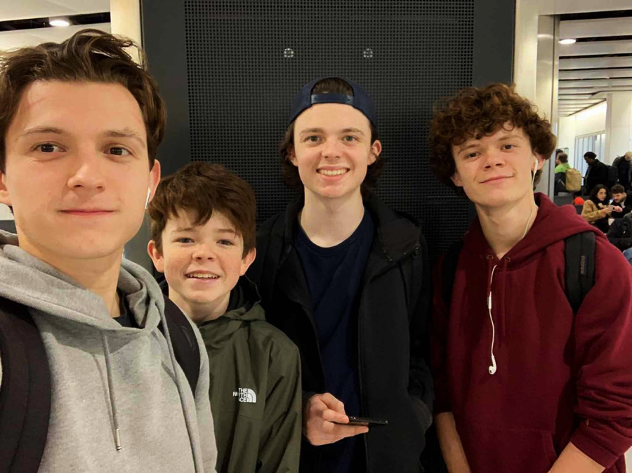 Four teenage boys are standing closely together indoors, smiling at the camera. Three wear hoodies and backpacks, one wears a cap backwards. There are other people and luggage in the background, suggesting an airport or station.