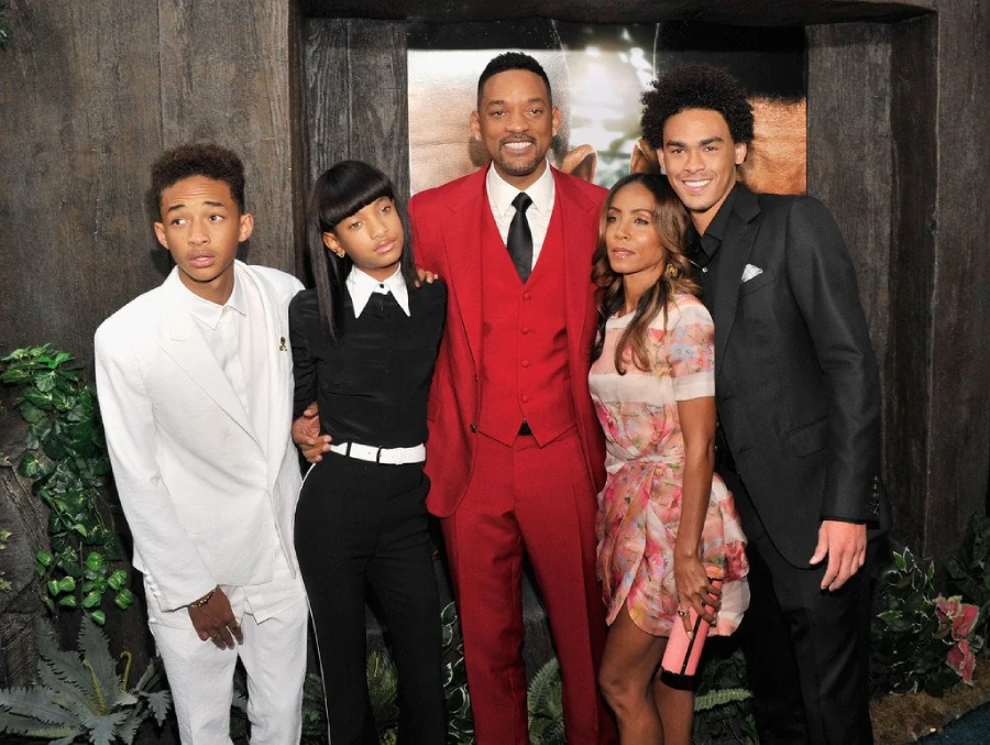 Five people posing together and smiling. The man in the center wears a red suit, flanked by two young people in black and white outfits, and a woman and another young man in a floral dress and black suit, respectively.