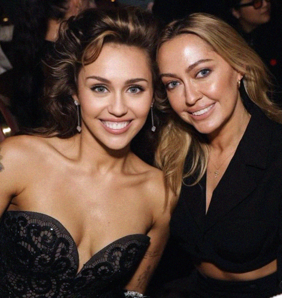 Two women smiling and posing together at an indoor event. One is wearing a strapless black lace dress, and the other is in a black outfit with long sleeves. Both have long hair and are looking directly at the camera.