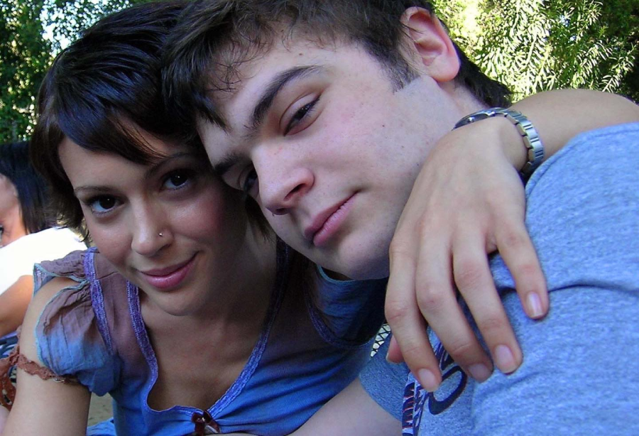 Two young people sit close together outdoors, smiling slightly at the camera. The person on the left has short hair and a nose ring, and their arm is draped around the person on the right, who has light skin and short brown hair.