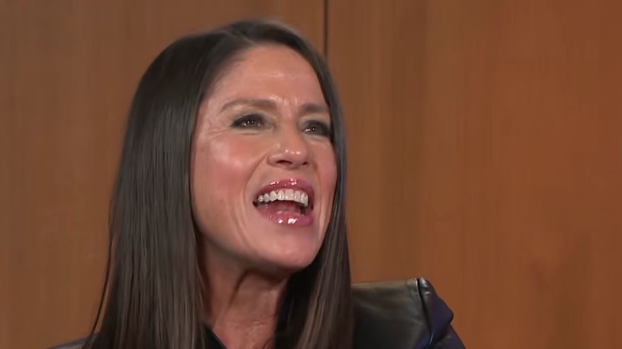 A woman with long brown hair is smiling and talking. She is wearing a dark top and is seated in front of a wooden background.