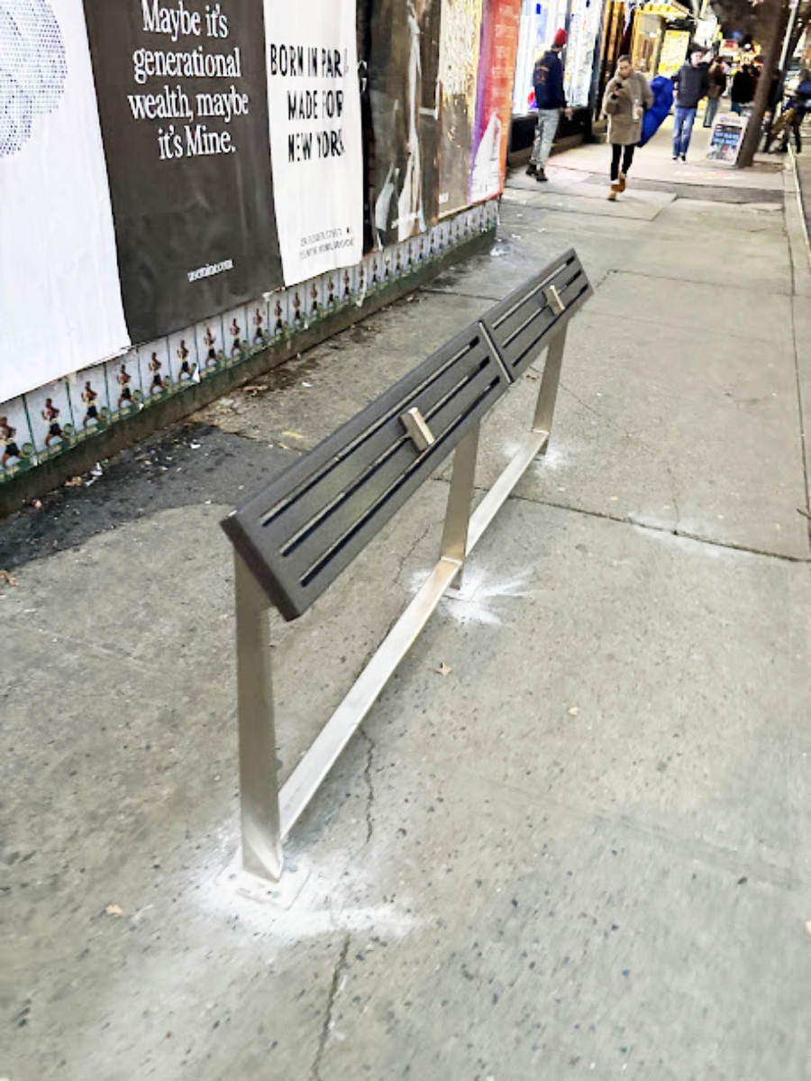 A slanted metal bench with divided seats is installed on a city sidewalk. People and colorful posters are visible in the background along a busy street.