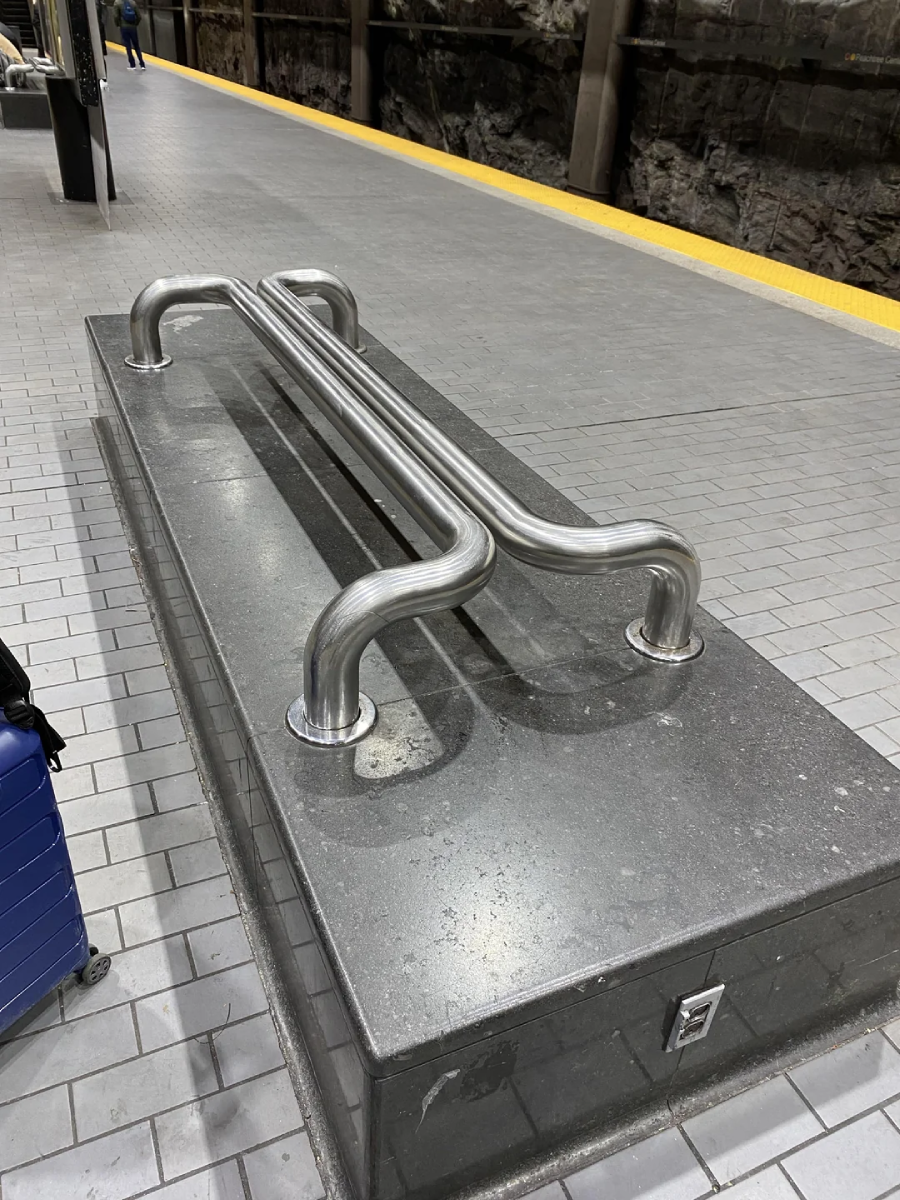 A metallic bench with curved bars instead of a flat seat, placed on a stone platform in a subway or train station. A blue suitcase sits nearby on the tiled floor; yellow tactile paving marks the platform edge.