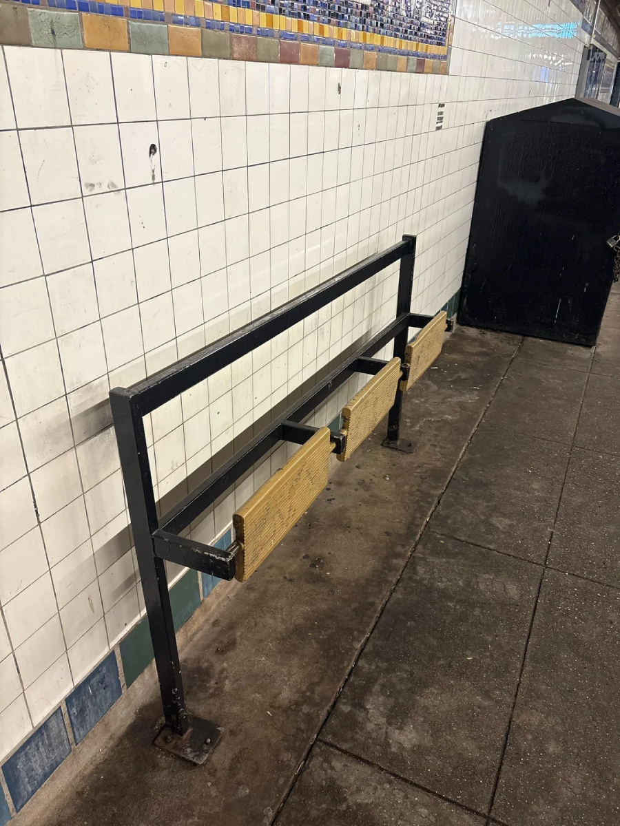 A metal bench frame with three separated wooden seat panels and no backrest is mounted against a tiled subway station wall. The floor shows dirt and stains, and a large black trash bin is nearby.