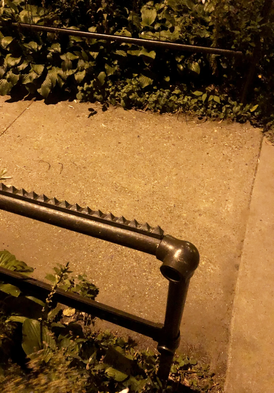 A metal railing with sharp triangular spikes lines a concrete pathway at night, surrounded by leafy green plants and partially illuminated by yellowish street lighting.