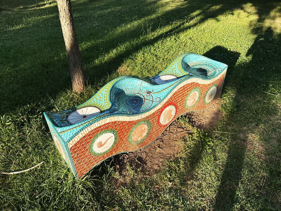 A wavy, colorful mosaic bench with circular designs sits on grass under a tree, casting shadows on the ground. The bench has blue, red, and orange tiles and text along its side.