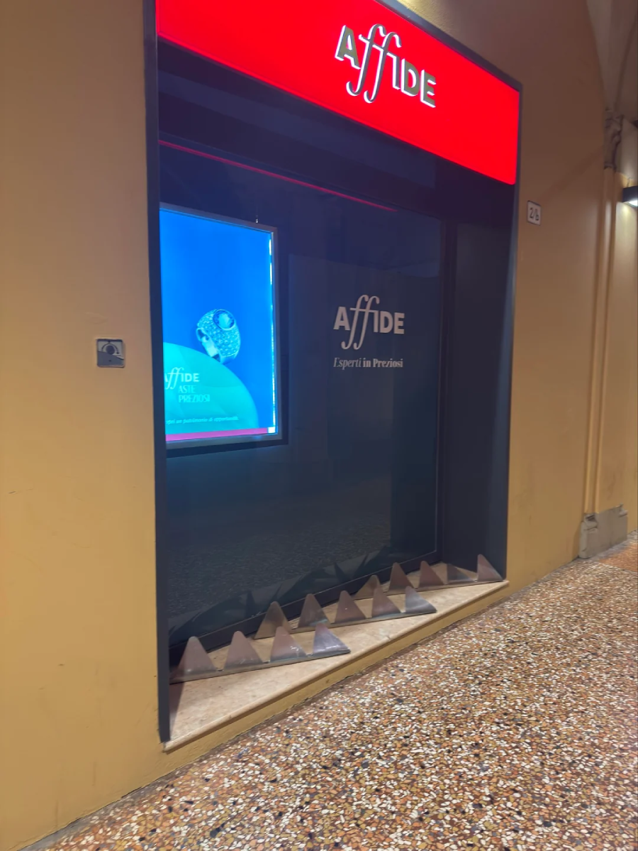 A shop window with a bright red "Affide" sign above and a display screen inside. The store entrance is blocked by a row of metal spikes on the ground. The floor is a speckled brown and beige terrazzo.