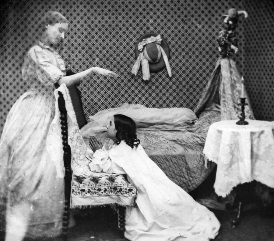 A vintage black-and-white photo shows a woman standing by a bed with her hand raised, while a girl kneels in prayer. The room has patterned wallpaper, a small table with a candle, and a hat hanging on the wall.