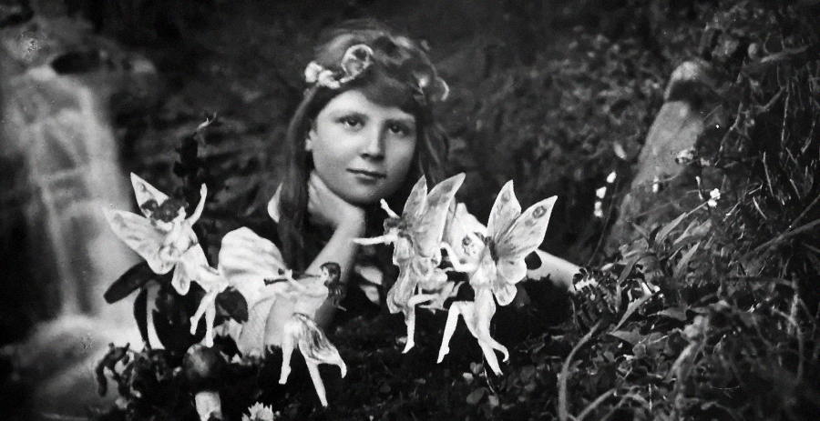 A vintage black-and-white photo of a young girl with long hair and a flower crown, resting her chin on her hand, surrounded by illustrated paper fairies in a garden setting with plants and a waterfall in the background.