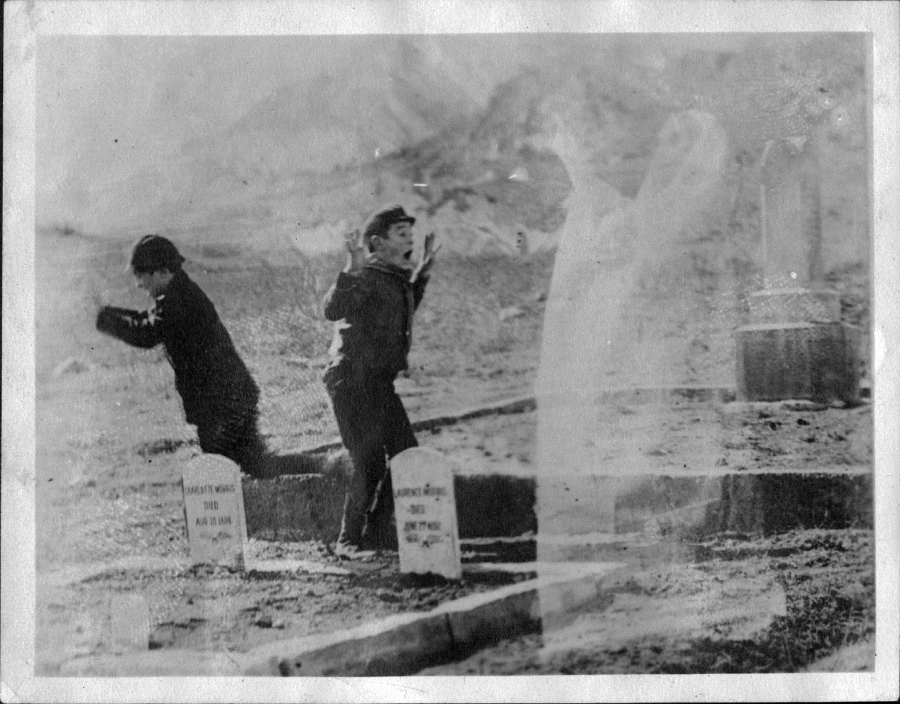 Two people appear to flee in fear near gravestones in a cemetery, while a ghostly, translucent figure stands between them, giving the impression of a supernatural encounter. The image is black and white and slightly blurry.