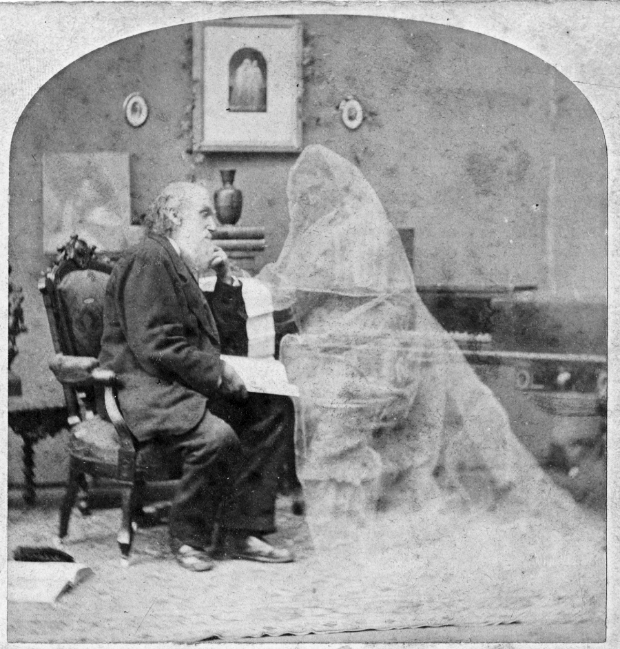 A sepia-toned vintage photograph shows a man sitting on a chair indoors, facing a faint, ghostly figure draped in translucent fabric, creating a supernatural effect. Paintings and furniture are visible in the background.