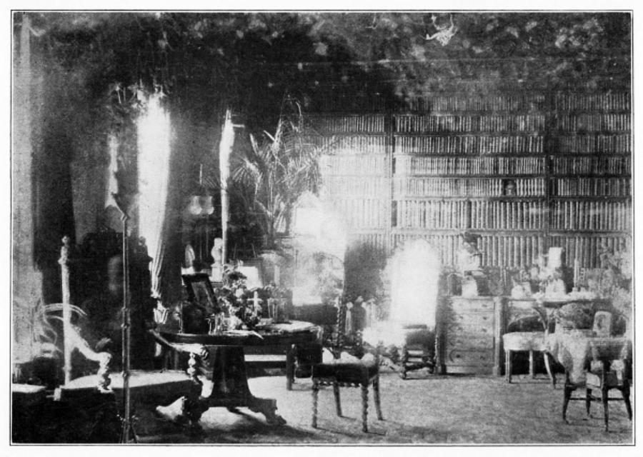 A black-and-white photo of a vintage library room, filled with bookshelves, tables, chairs, and ornate decor. Bright light from windows creates reflections and bright spots throughout the image.