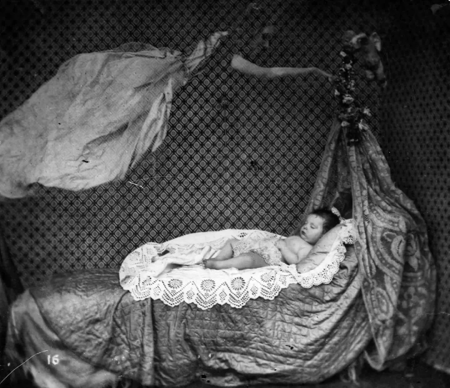 A black and white photo of a baby sleeping on a bed draped with lace and fabric. Above, a translucent, ghostly figure appears to float toward the baby in a patterned, Victorian-style setting.