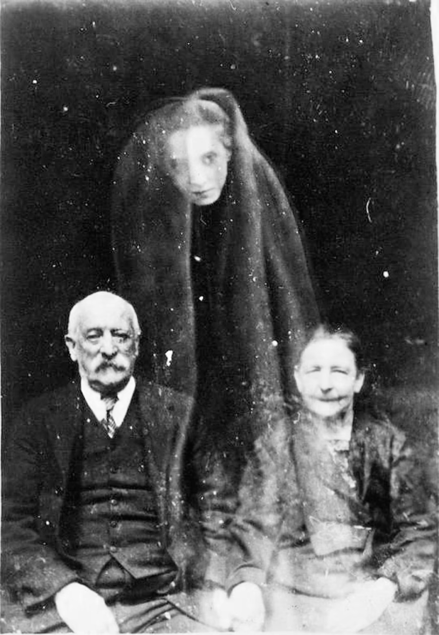A vintage black-and-white photo shows an elderly man and woman seated, with a transparent, ghostly figure of a young woman draped in a veil standing behind them.
