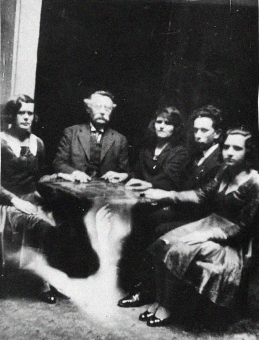 Five people sit around a table with their hands resting on it; a blurry, ghostly shape appears beneath the table in the foreground, creating an eerie atmosphere.
