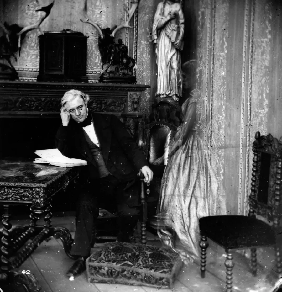 A man in 19th-century clothing sits at an ornate desk, leaning his head on his hand and looking thoughtful. Papers lie on the desk, and a statue and elaborate decorations are visible in the background.
