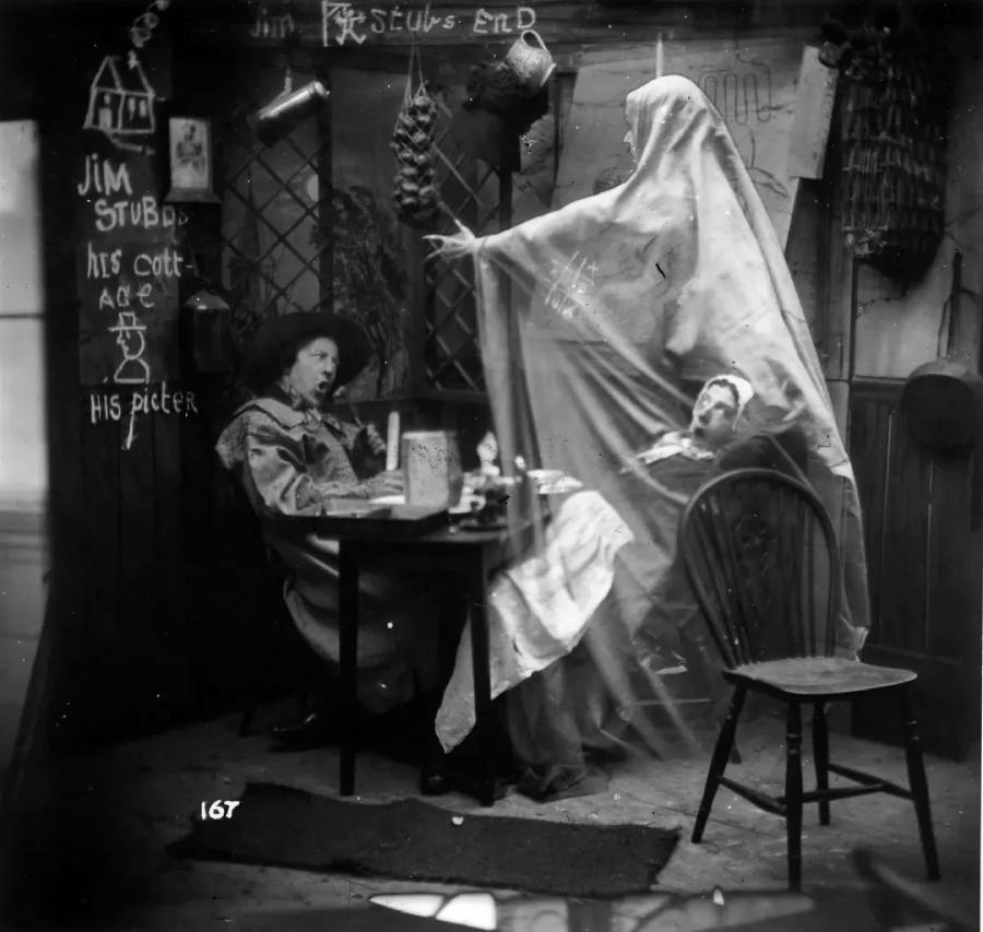 Two people sit at a table in a cluttered room as a figure draped in a ghostly white sheet looms over them with an outstretched arm. The walls are decorated with chalk drawings and writing.