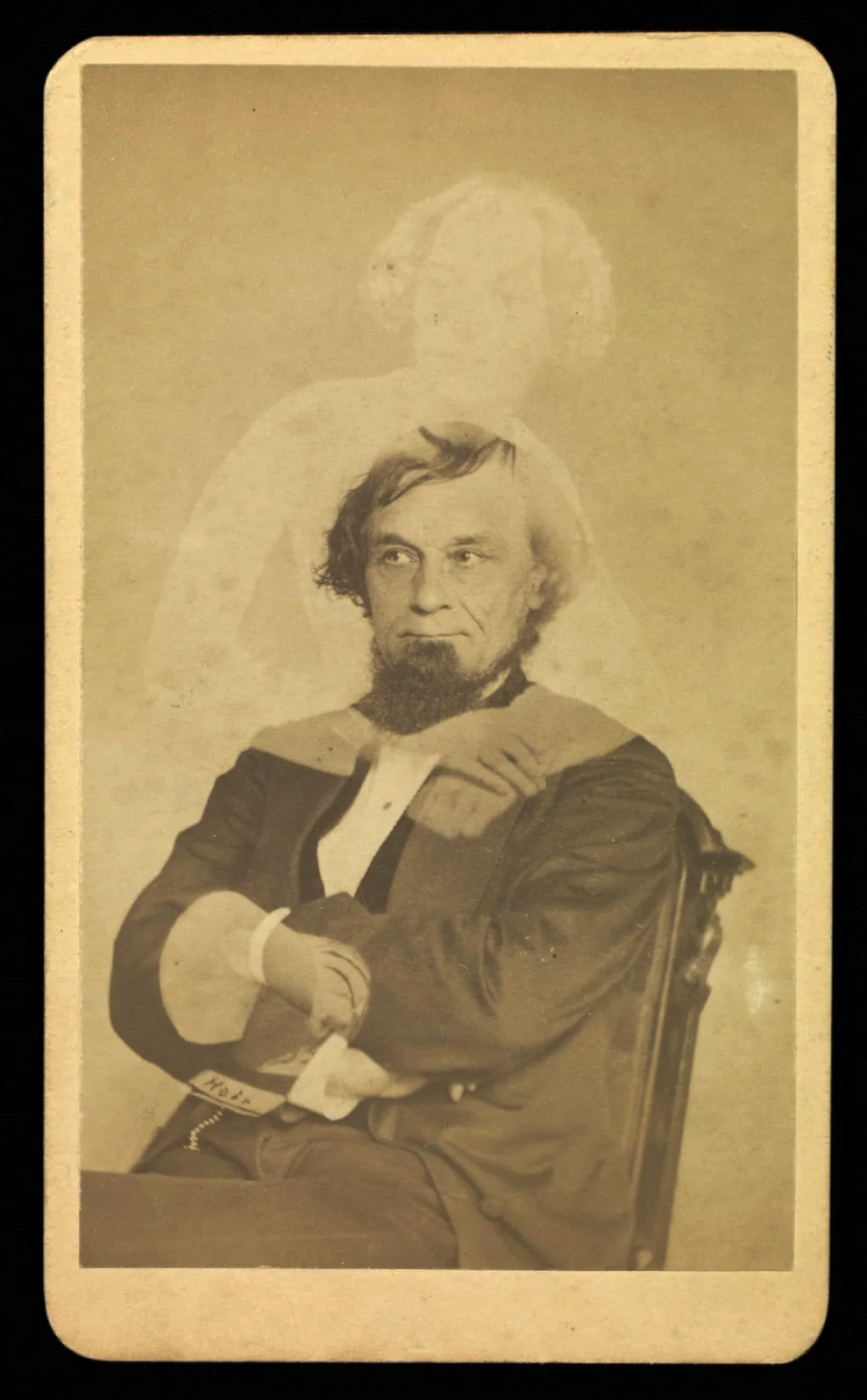 A sepia photograph of a seated man with a beard, dressed in 19th-century clothing, with a faint ghostly figure of a woman appearing to rest her hands on his shoulders behind him.