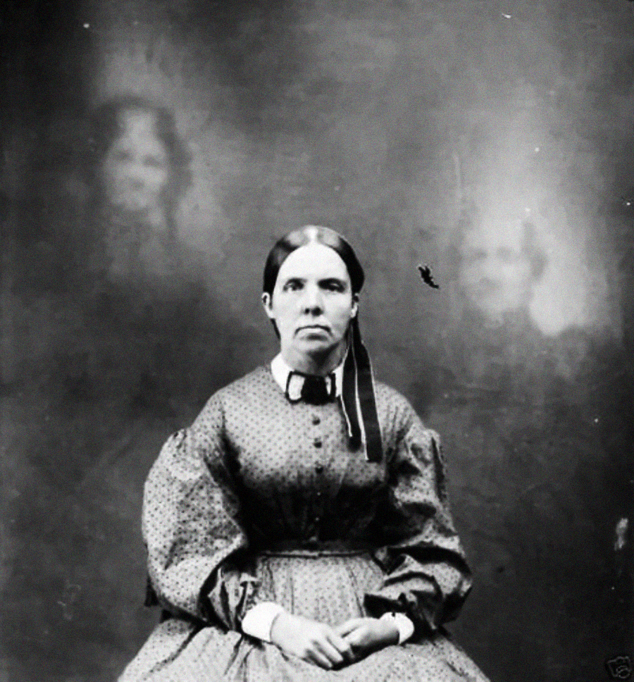 A woman in a patterned dress sits facing the camera, hands folded on her lap, with two faint, ghostly figures visible in the hazy background behind her. The photo is black and white and appears old.